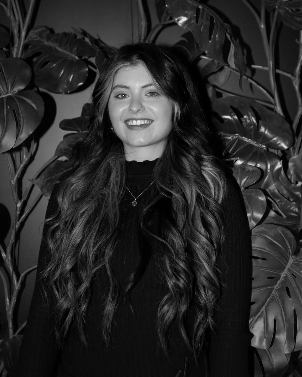A woman with long wavy hair smiling, standing in front of large leafy plants.