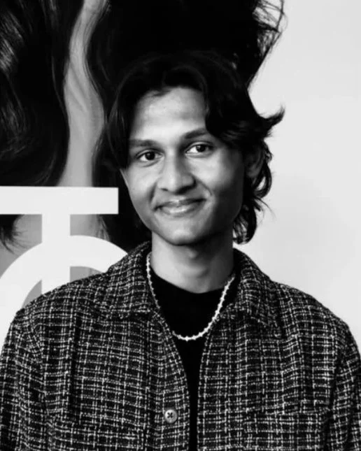Black and white photo of a young man with wavy hair wearing a checkered jacket, a black shirt, and a pearl necklace, smiling at the camera.