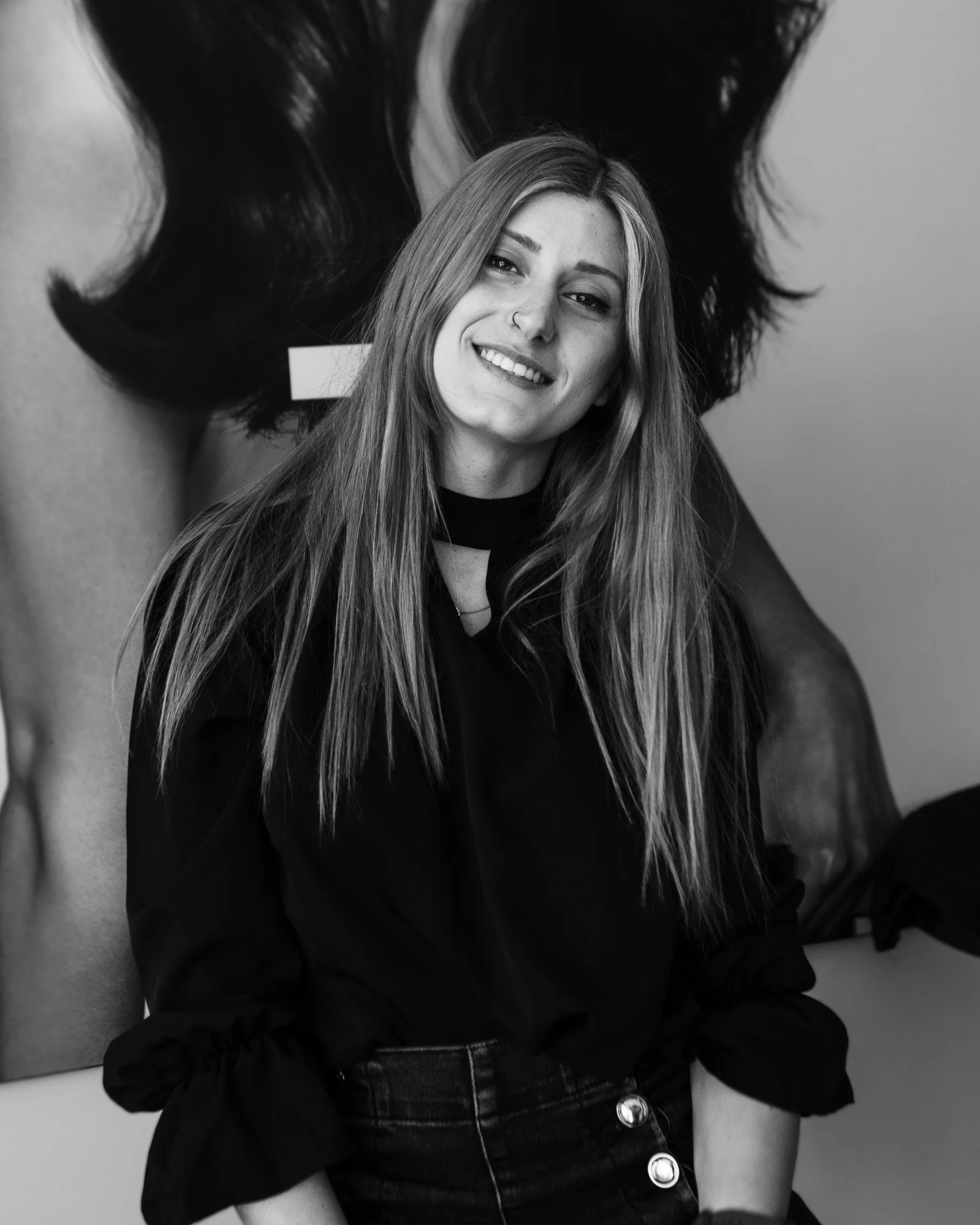 Black-and-white photo of a young woman with long hair, smiling, wearing a dark top with rolled-up sleeves and high-waisted jeans, standing in front of an artwork.