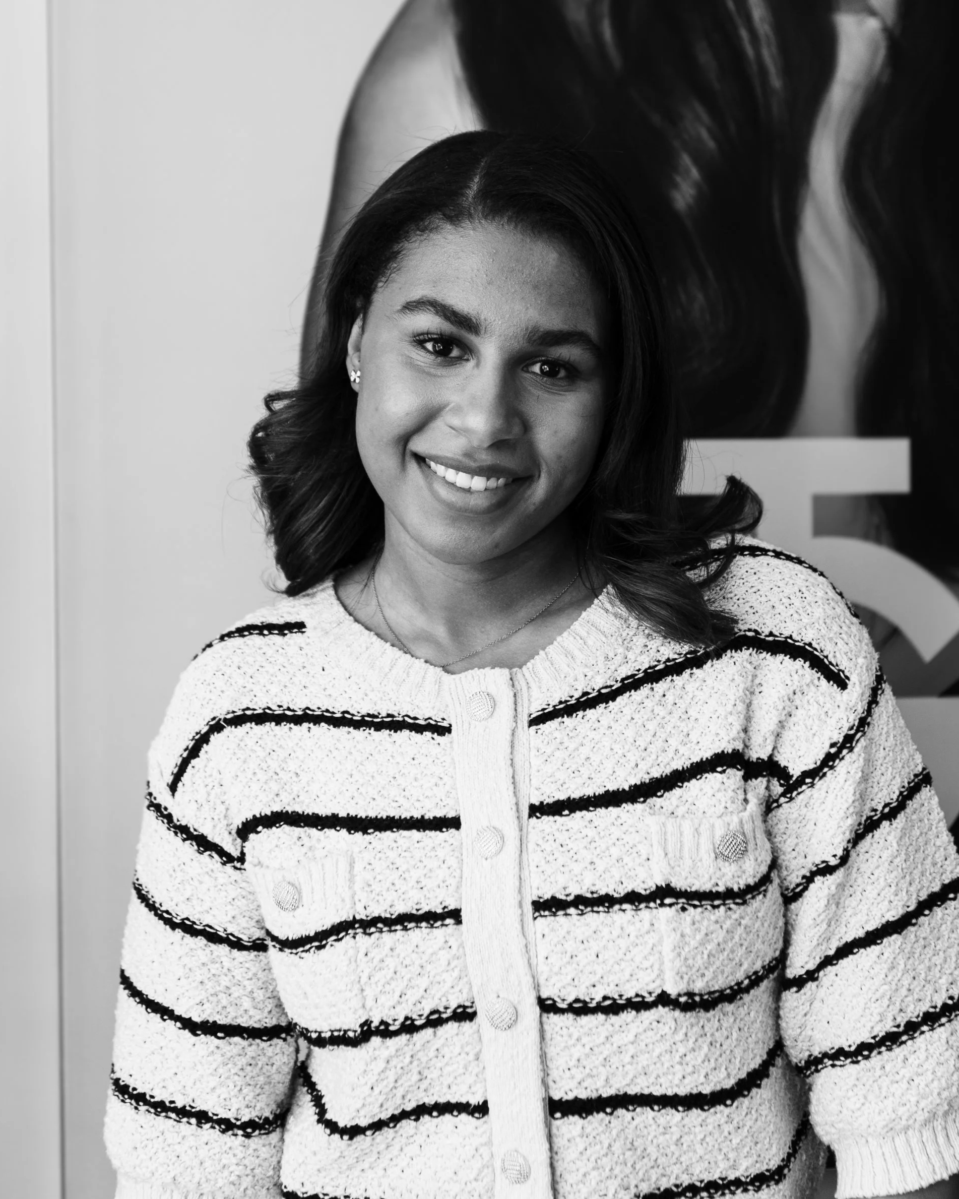 A smiling woman with dark hair, wearing a striped knit sweater, standing in front of a poster.