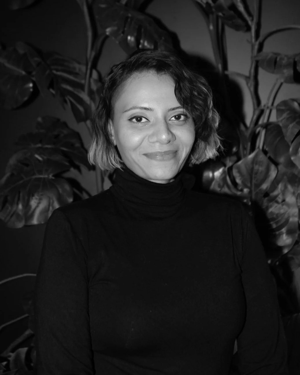 A black and white portrait of a woman with short, wavy hair, wearing a turtleneck, standing in front of large leafy plants.
