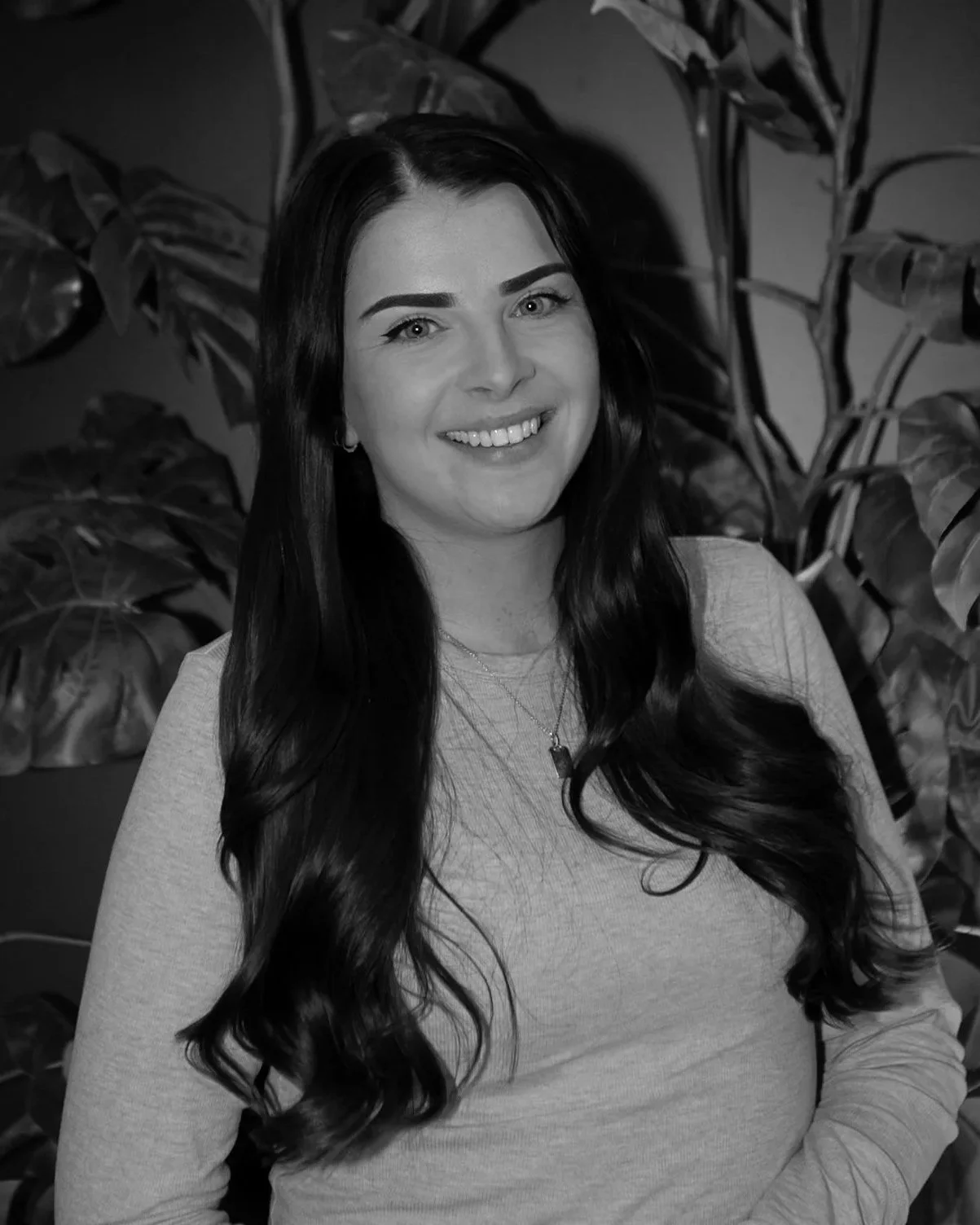 Black and white portrait of a smiling woman with long dark hair, wearing a light-colored top, standing in front of large leafy plants.