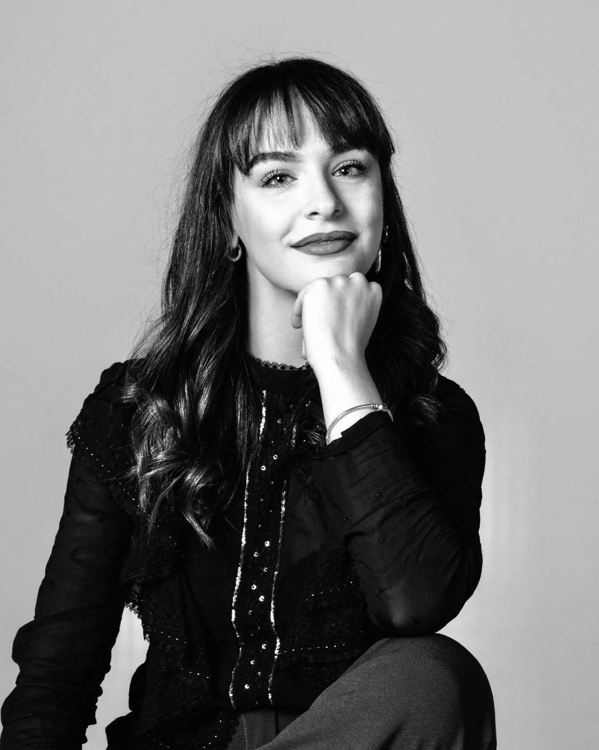 Black and white portrait of a woman with long wavy hair and bangs, smiling, with her chin resting on her hand, against a plain background.