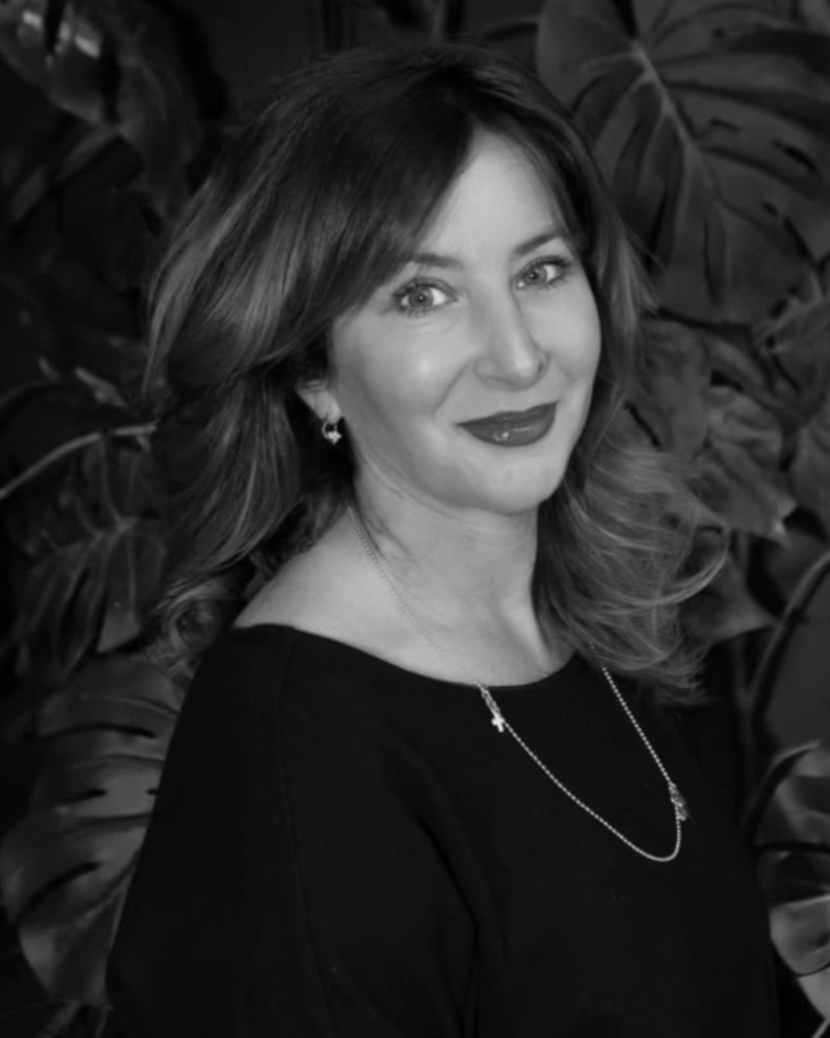A woman with shoulder-length wavy hair, wearing earrings, a necklace with a cross, and a dark top, smiling in front of large tropical leaves.