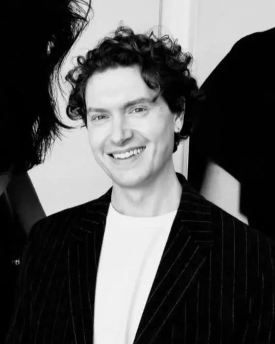 A smiling man with curly hair wearing a pinstripe blazer and white shirt, surrounded by partially visible people.
