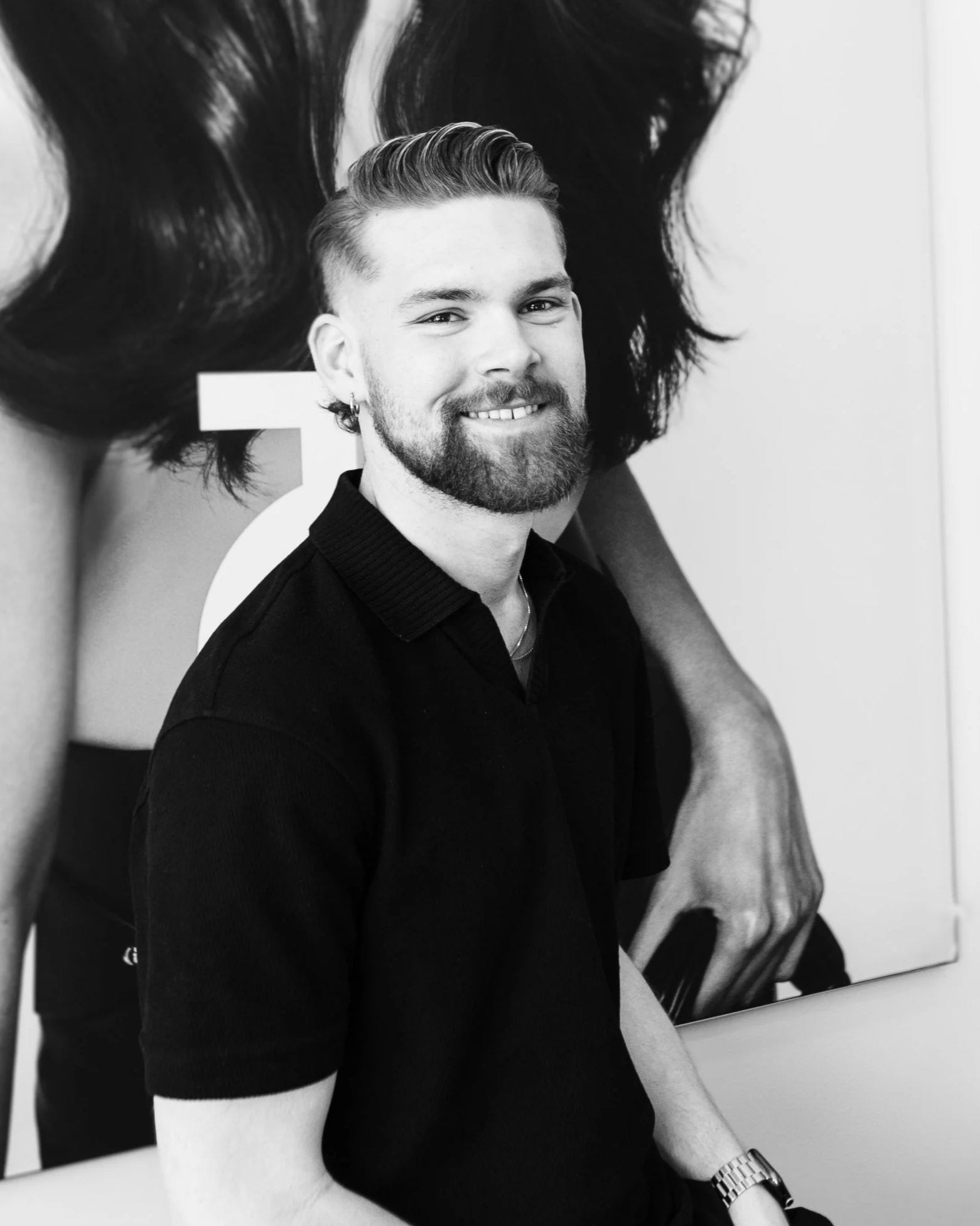 A young man with a beard and short hair smiling, sitting in front of a large photograph of a woman with long wavy hair.