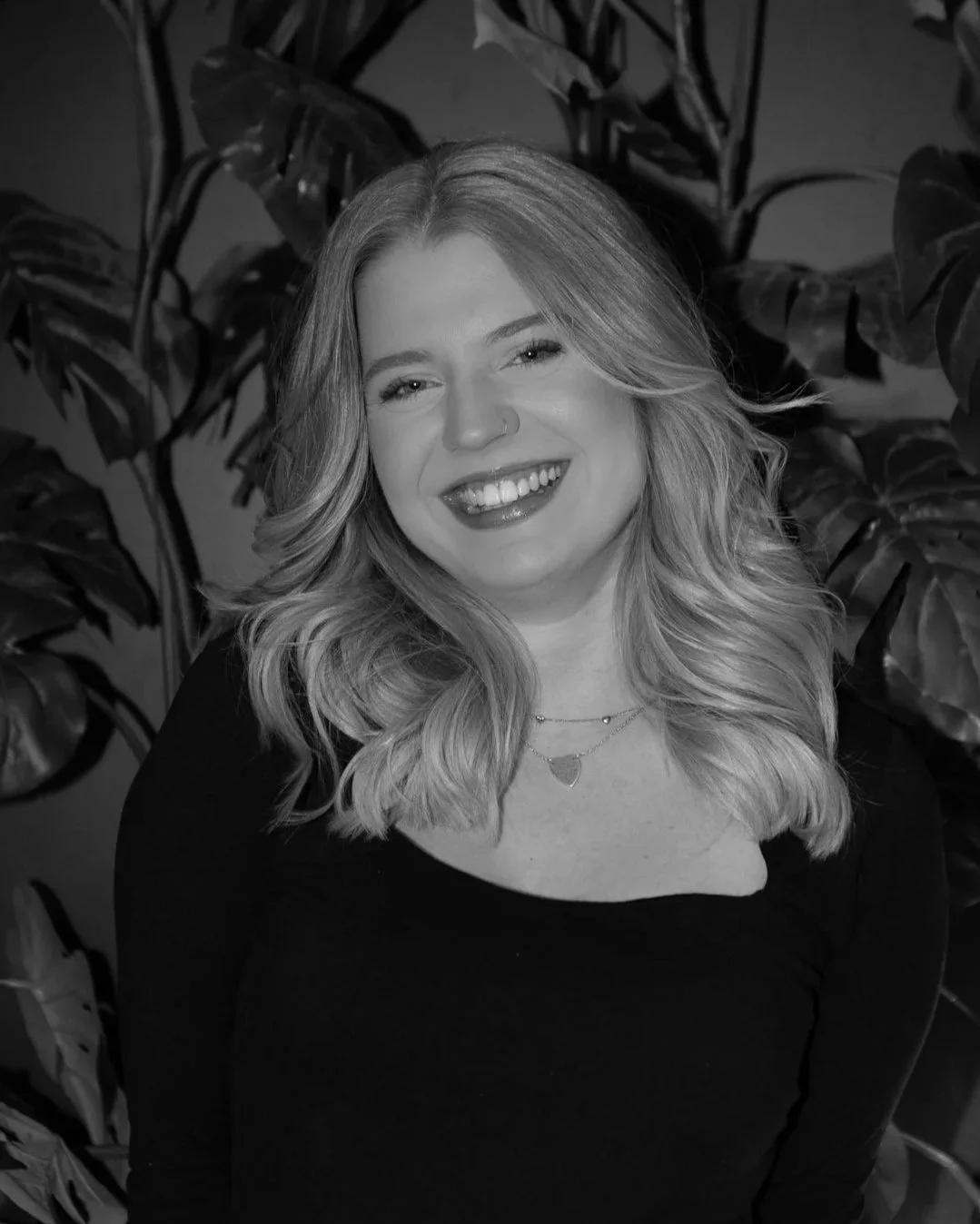 Black and white portrait of a young woman with wavy blonde hair, smiling, wearing a black top, jewelry, and standing in front of leafy plants.