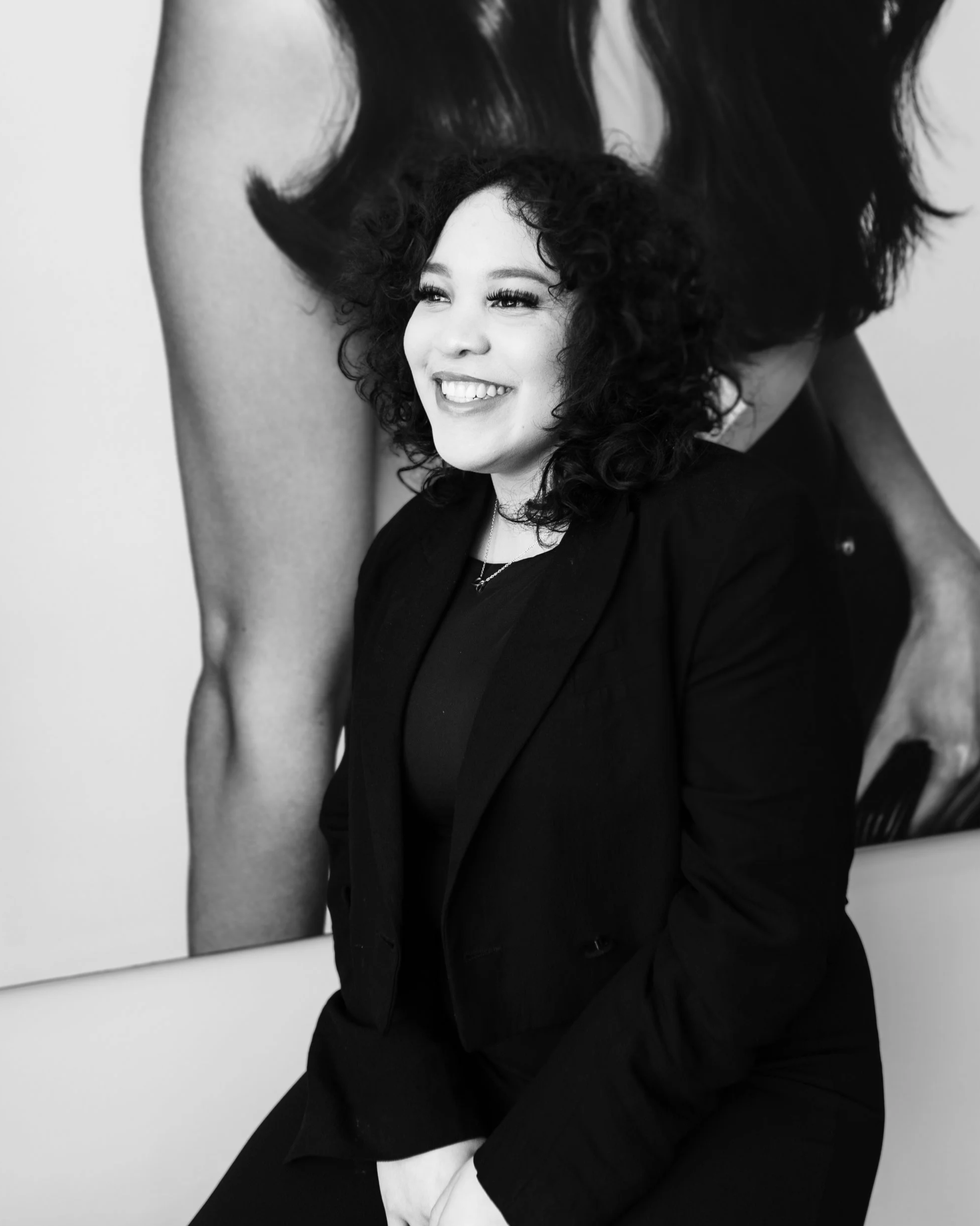 A woman with curly hair smiling, dressed in black, sitting in front of a large black and white portrait of a woman with long hair.