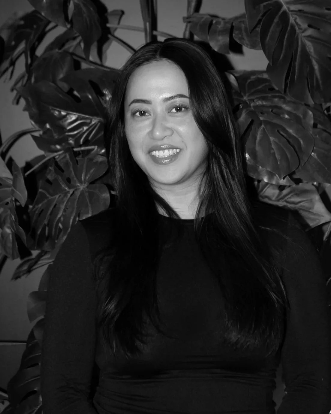 A woman with long dark hair smiling, standing in front of large leafy plants.