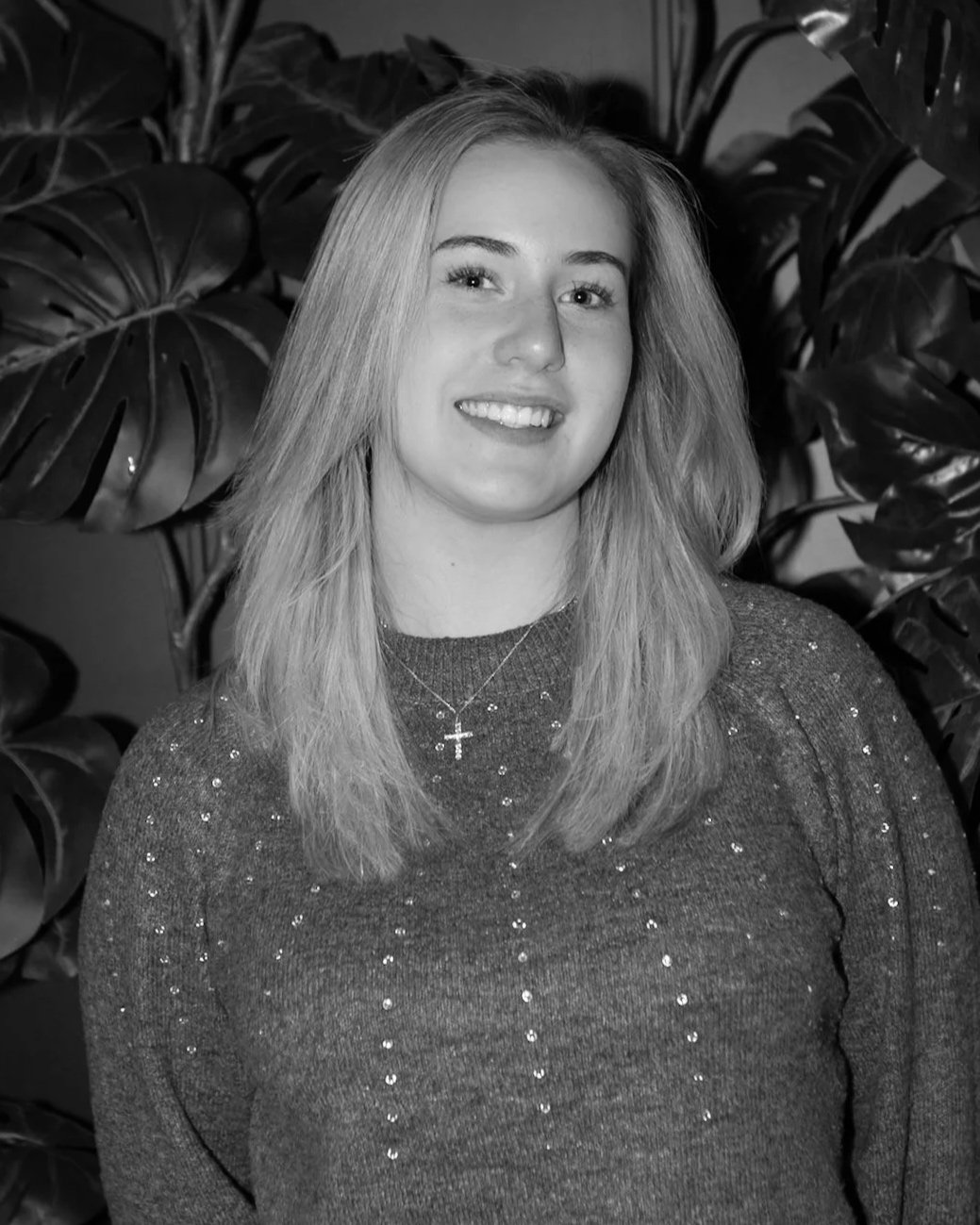 A young woman with shoulder-length blonde hair smiling at the camera, wearing a dark, sparkly sweater with a cross necklace, standing in front of large tropical leaves.
