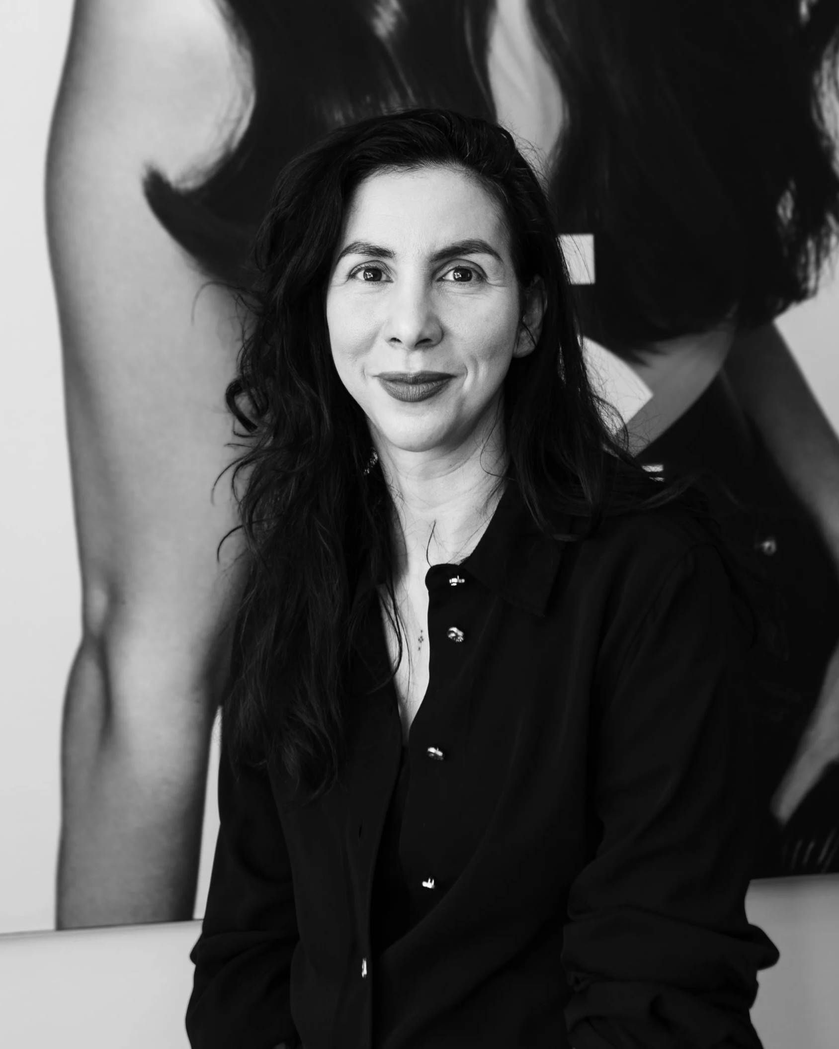 Black-and-white portrait of a woman with long dark hair, smiling, wearing a dark button-up shirt, sitting in front of an abstract artwork featuring a partially visible figure with dark hair.