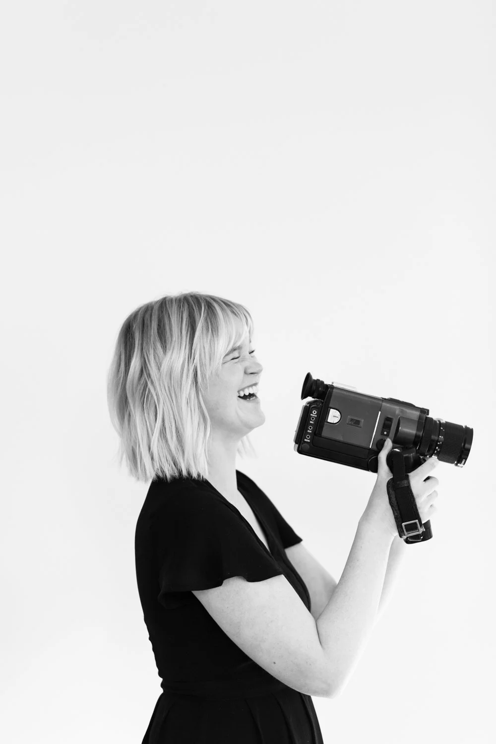 Black and white photo of a woman with short blonde hair laughing while holding a vintage camera.