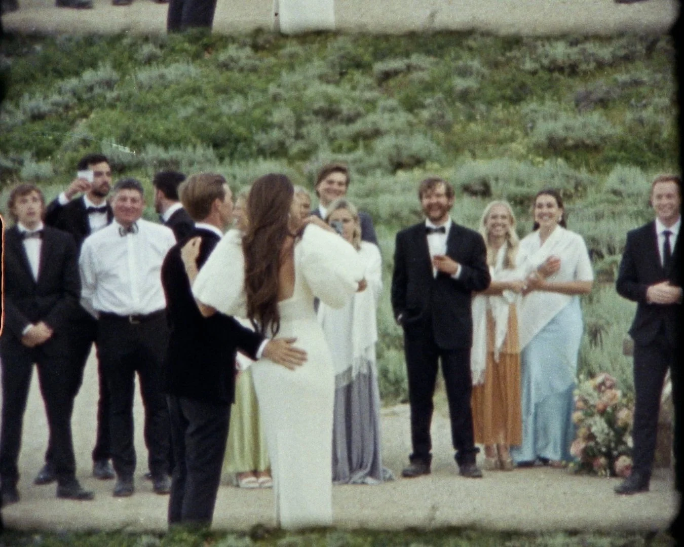 A wedding celebration outdoors with a bride and groom embracing, surrounded by friends and family in formal attire, with a lush green hillside in the background.