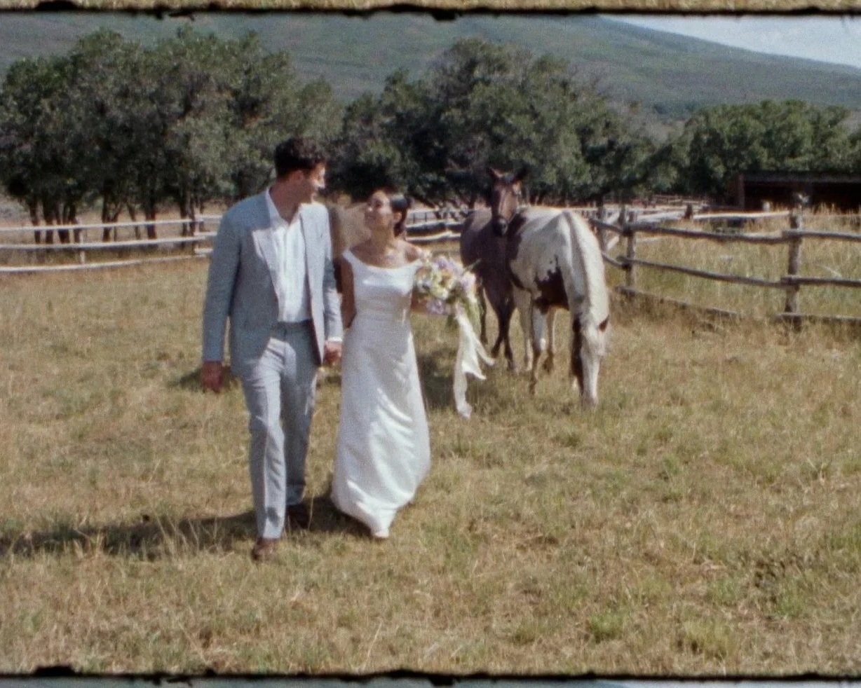 A bride and groom holding hands and walking in a grassy outdoor area, with a white and brown horse nearby, surrounded by fencing and trees, during daytime.