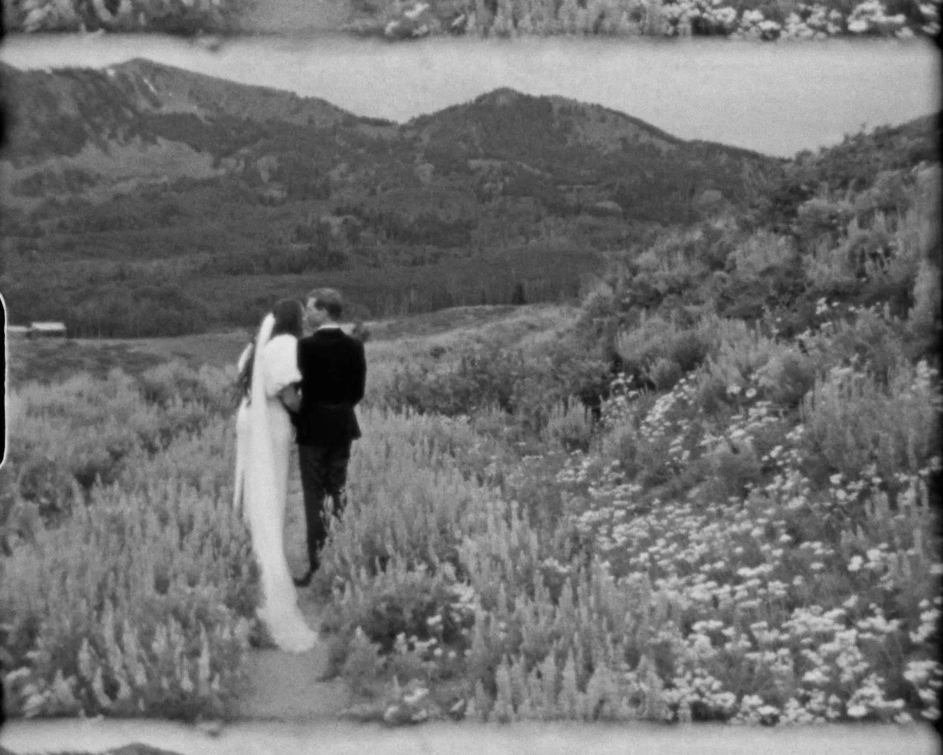Black and white photo of a bride and groom standing closely together in a field of flowers, with hills and a lake in the background.