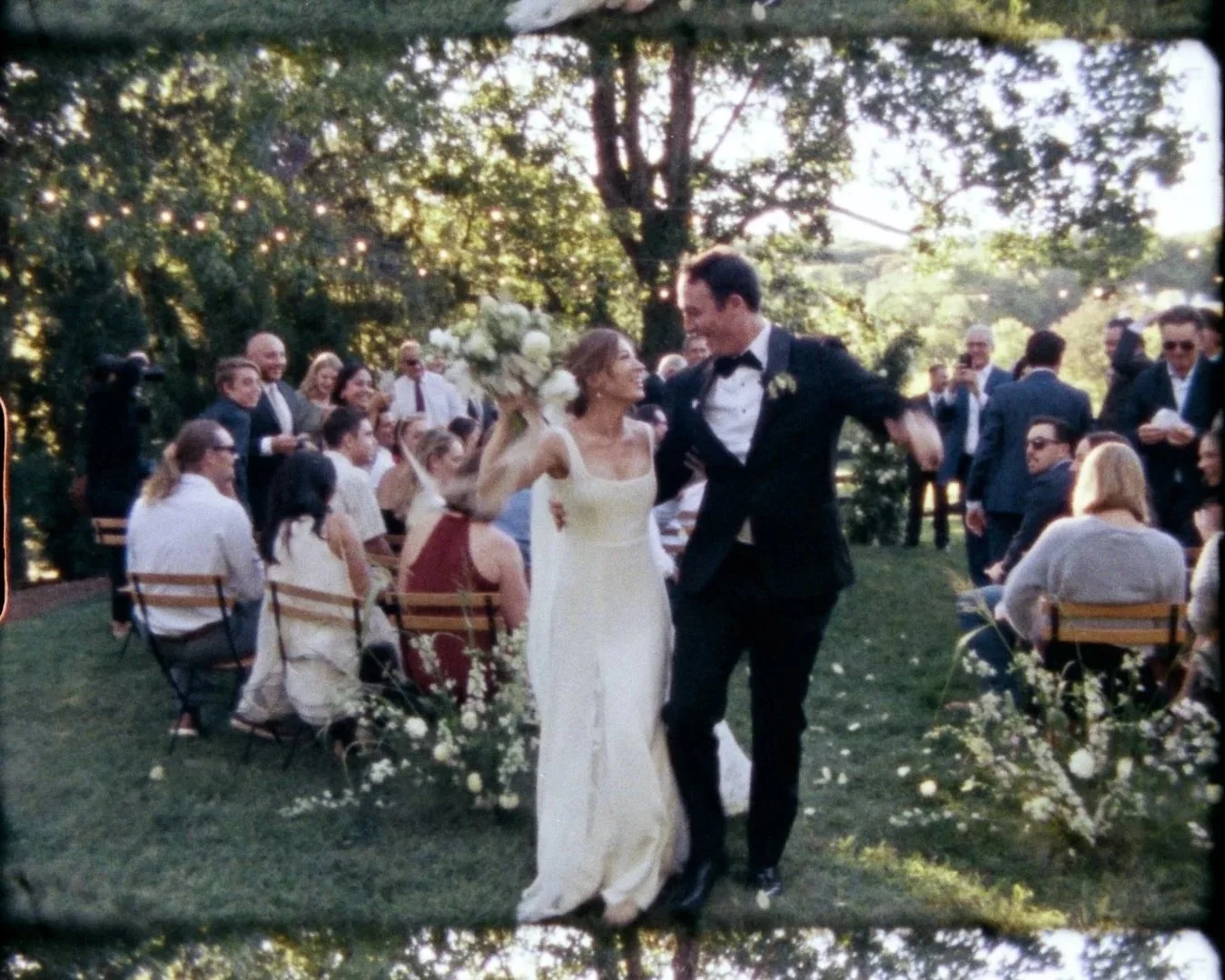 A bride and groom dancing outdoors at their wedding reception, surrounded by seated guests and standing onlookers in a garden setting with trees and string lights.
