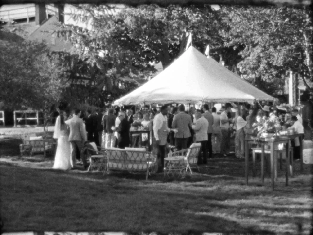 Black-and-white photo of a wedding reception under a large tent with guests mingling, some in formal attire, outdoors surrounded by trees and park furniture.