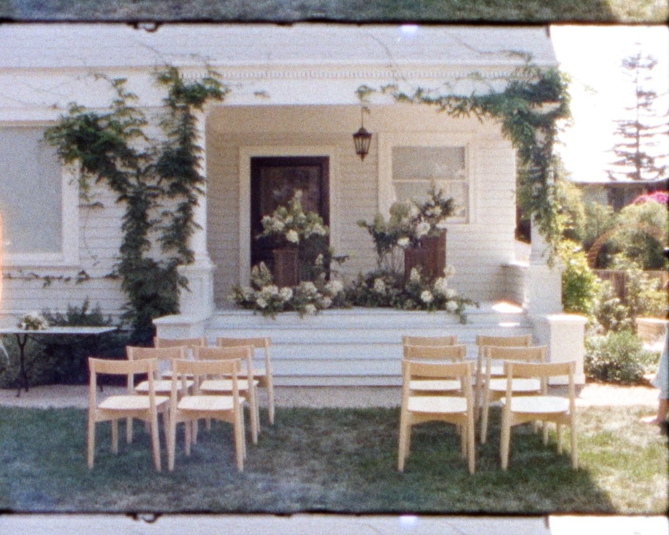 Outdoor wedding setup with wooden chairs facing a decorated porch with floral arrangements, ivy, and a hanging lantern.