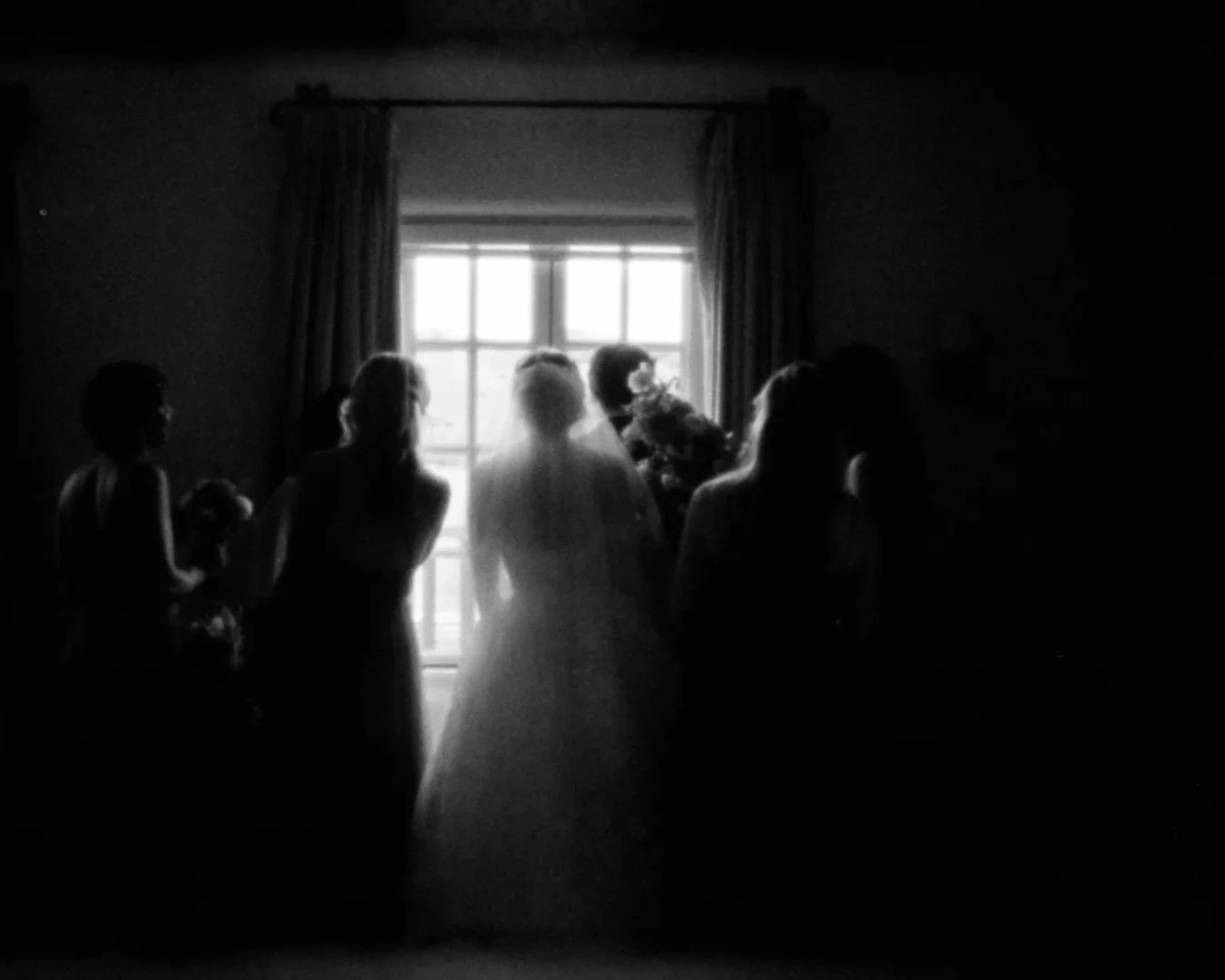 Silhouettes of people standing near a window with curtains, backlit by natural light, in a dimly lit room.