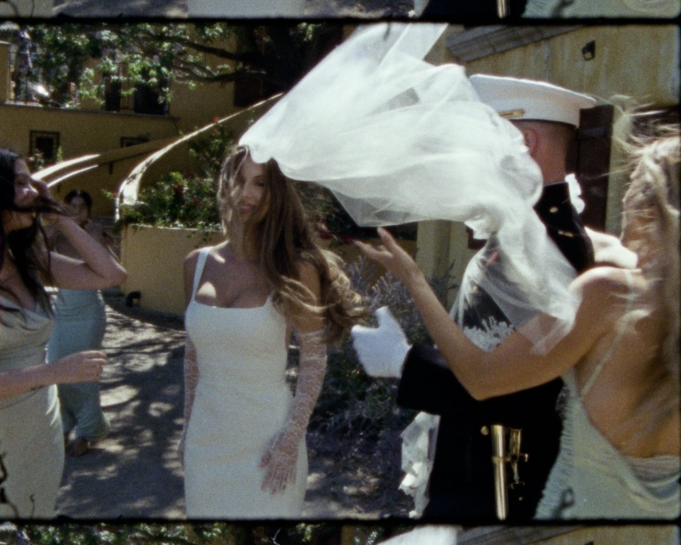 A woman in a white dress is getting her hair done with a hairspray during an outdoor event, surrounded by other women and a man in a uniform.