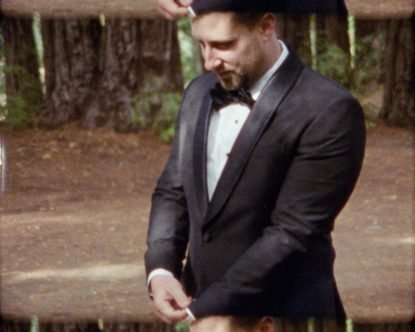 A man in a tuxedo adjusting his cufflinks in a wooded outdoor setting.