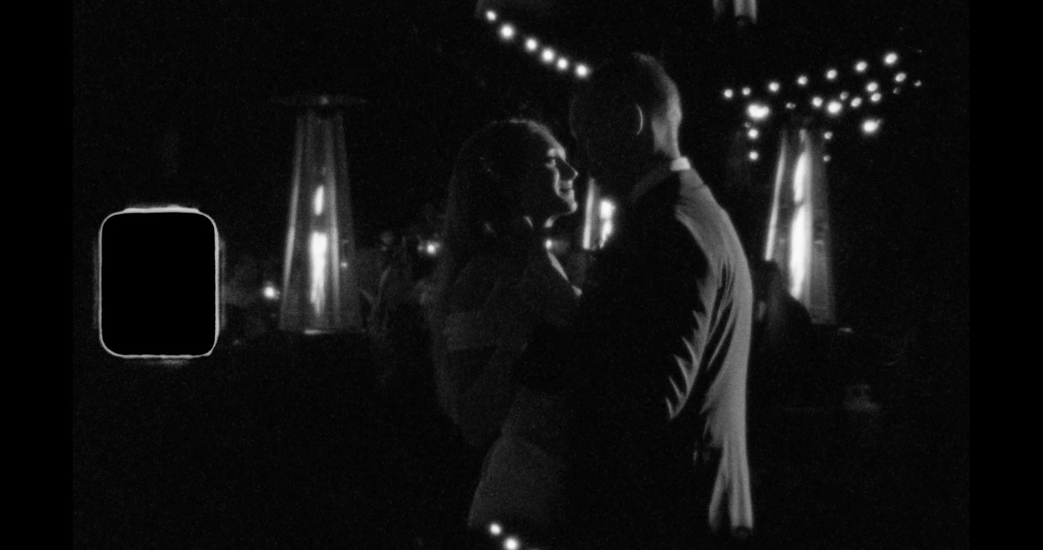 A black and white photo of a couple dancing closely at night, with string lights and tall lights in the background.