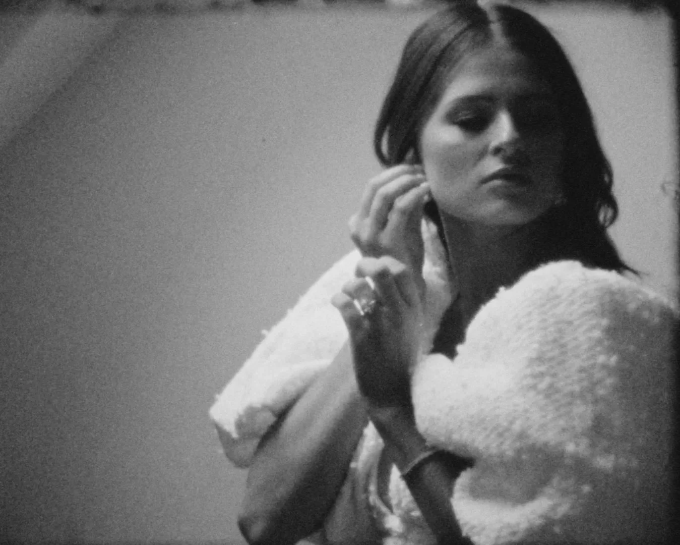 Black and white photo of a woman with medium-length dark hair, wearing a textured sweater, looking to her left with her hand near her ear.