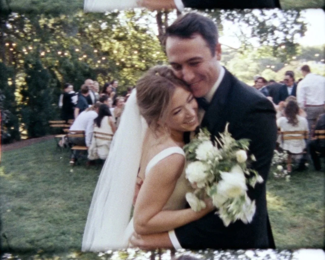 A newlywed couple hugging and smiling outdoors at their wedding reception, with guests seated at tables in the background and trees surrounding the area.