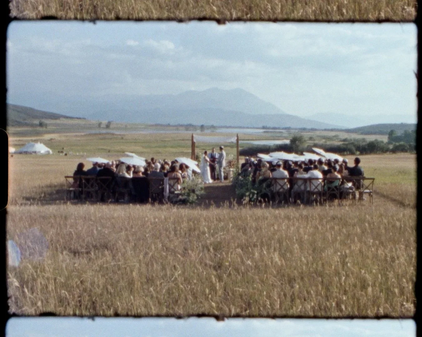Outdoor wedding ceremony with guests seated on benches under umbrellas, officiant and bride and groom at the altar, set in a scenic field with mountains in the background.