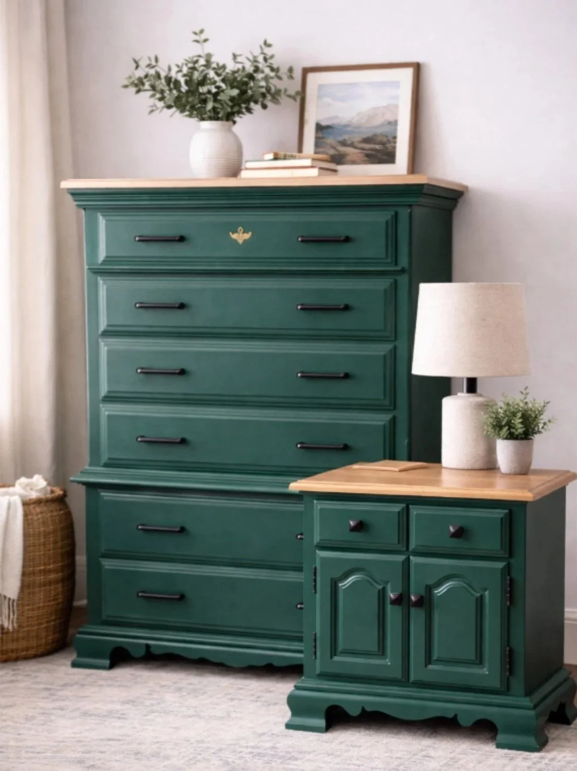 A green wooden dresser with five drawers and black handles, topped with a white vase with green foliage, a framed landscape painting, and a few books. A matching green wooden cabinet with two doors and black knobs, topped with a beige lamp and a small potted plant, is in front of the dresser. A basket with a towel is on the floor to the left.