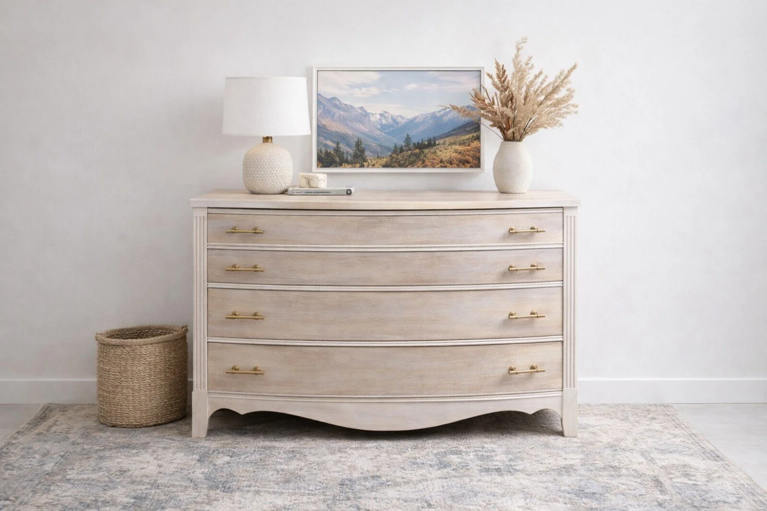 A white vintage dresser with five drawers and gold handles, topped with a white lamp, a stack of books, a framed mountain landscape photo, and a white vase with dried pampas grass; a woven basket is on the floor to the left.
