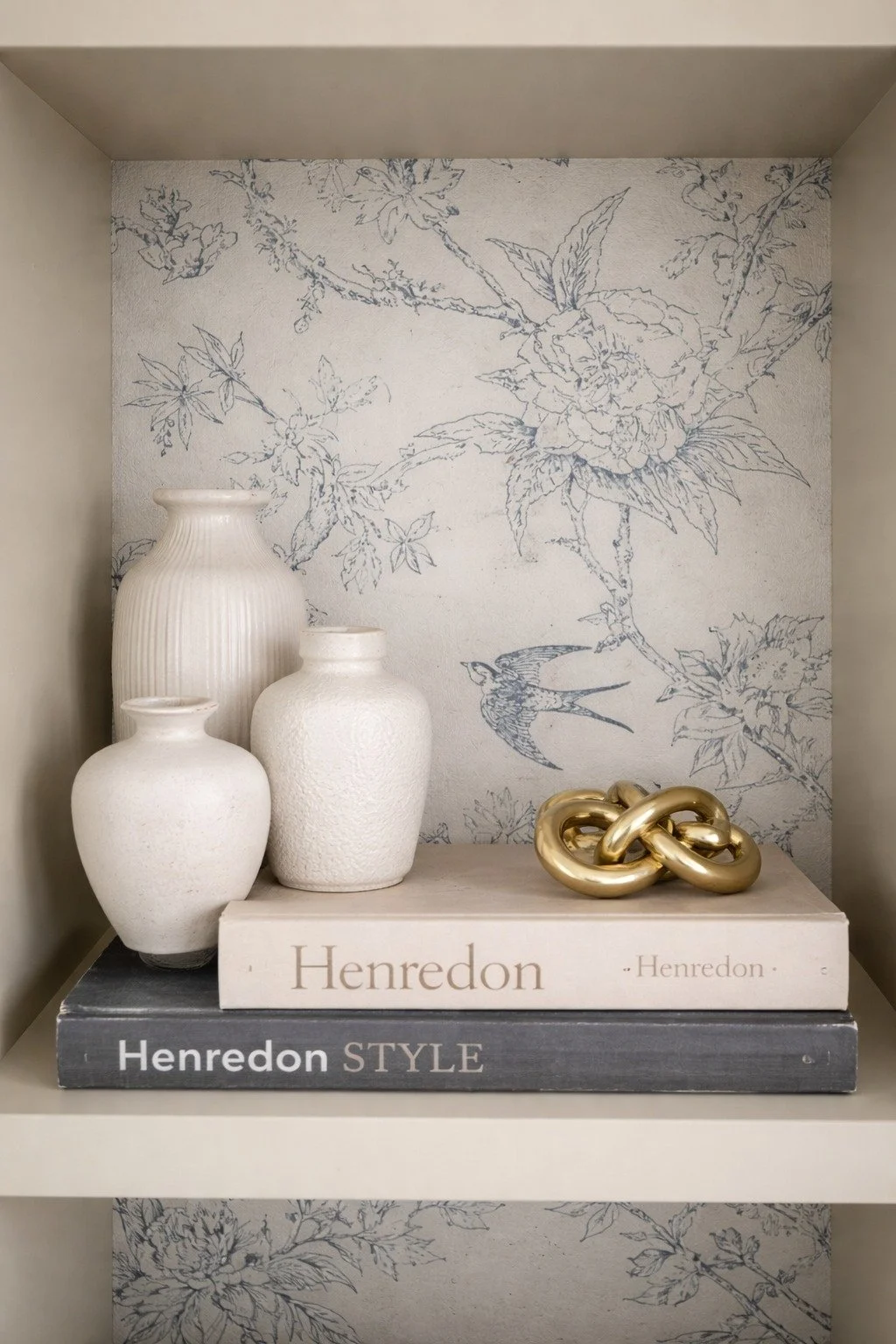 Decorative display with three white vases, a gold decorative knot, and books titled 'Henredon' and 'Henredon STYLE' on a shelf against a floral and bird wallpaper background.