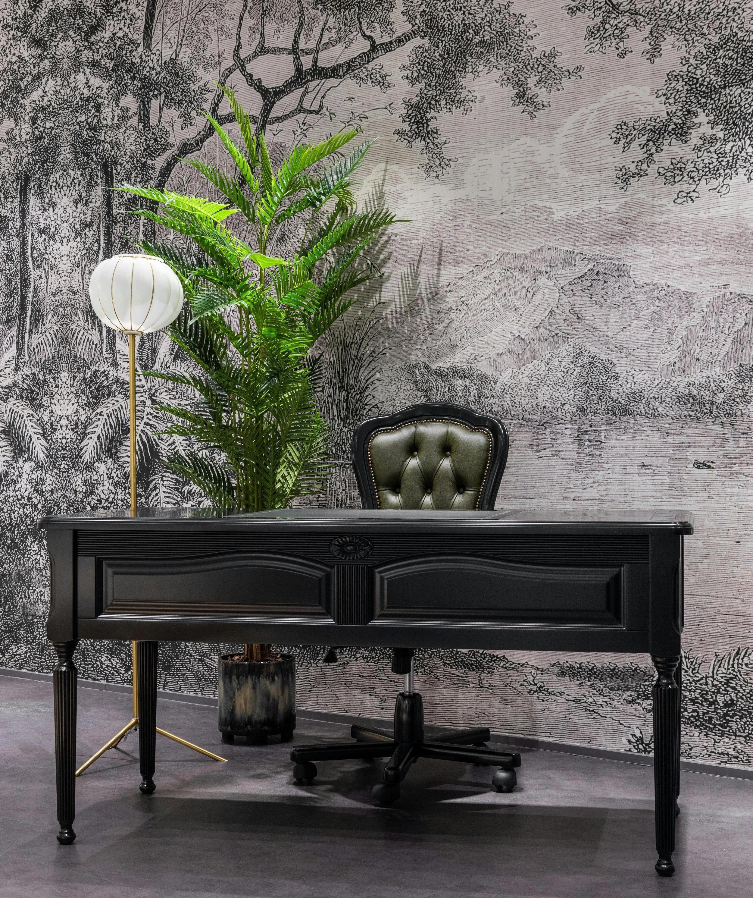 Office setup with a black wooden desk, green leather tufted chair, a tall green plant, a white standing lamp, and a scenic black and white mural wall featuring mountains and trees.