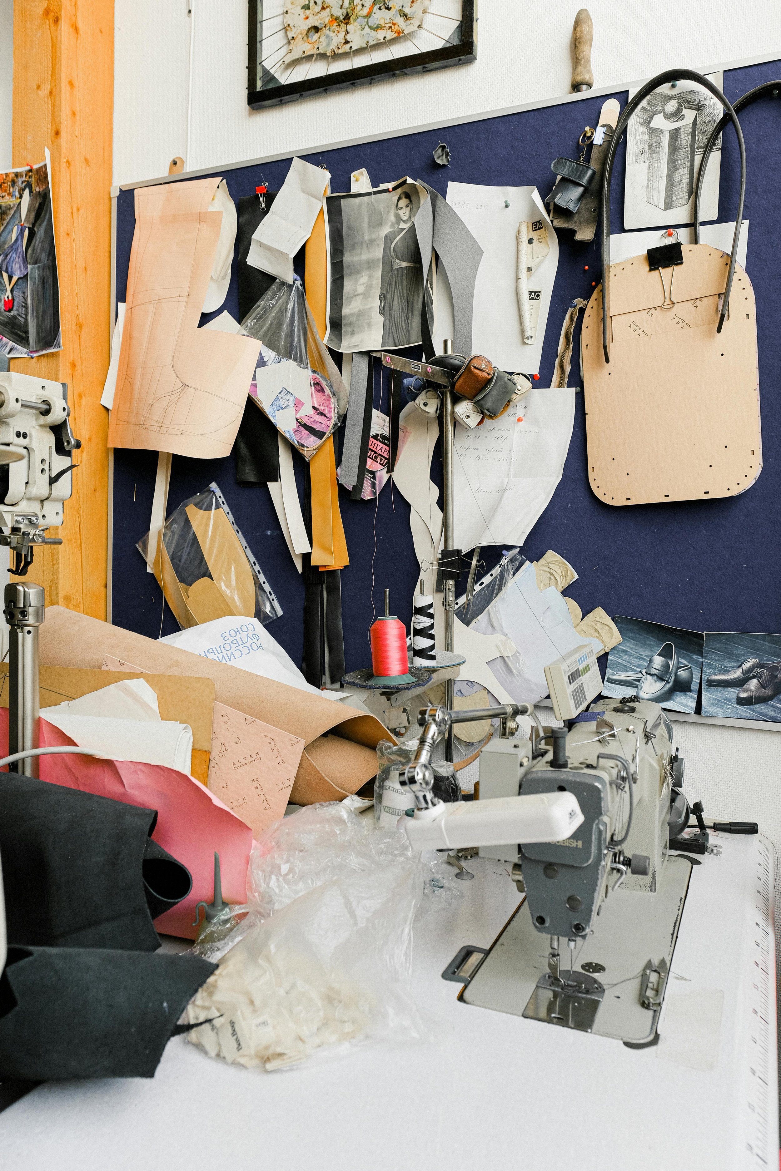 A sewing workspace with a sewing machine on a table, surrounded by fabric, patterns, threads, and design sketches, with a bulletin board filled with sketches and notes in the background.