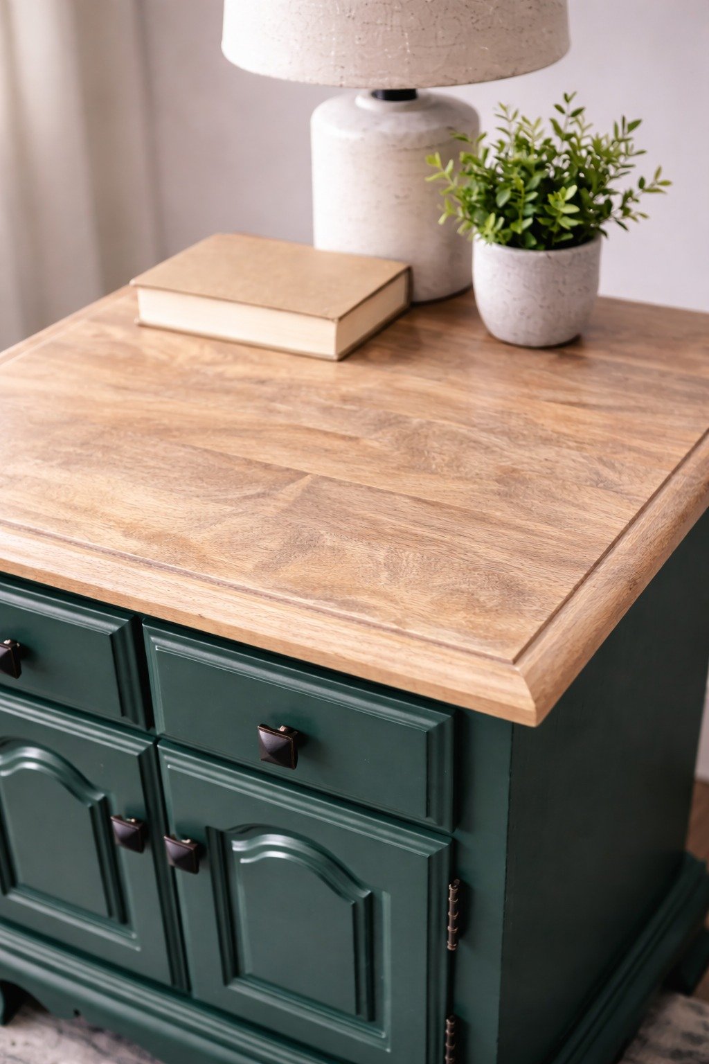 A wooden tabletop with a beige lamp, a beige book, and a small potted green plant on top. The table has dark green drawers and cabinet doors.