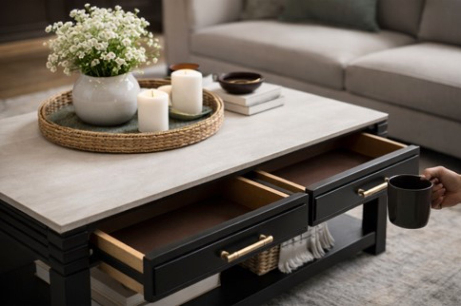 Coffee table with a white top, black legs, and gold handles, featuring a decorative tray with white candles, a white vase with white flowers, and books. A person's hand holding a black mug is visible as they place it on the table in a cozy living room setting.