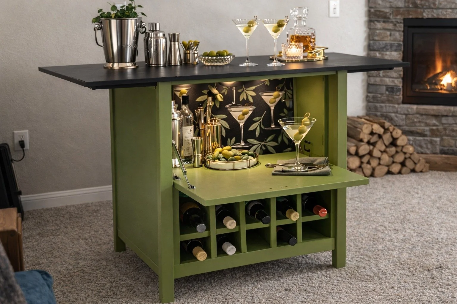 A green bar cart with a decorated back panel, stocked with bottles and bar accessories, topped with a black countertop featuring martini glasses with green olives, and a tray of green olives. In the background, a cozy fireplace and a pile of firewood are visible.
