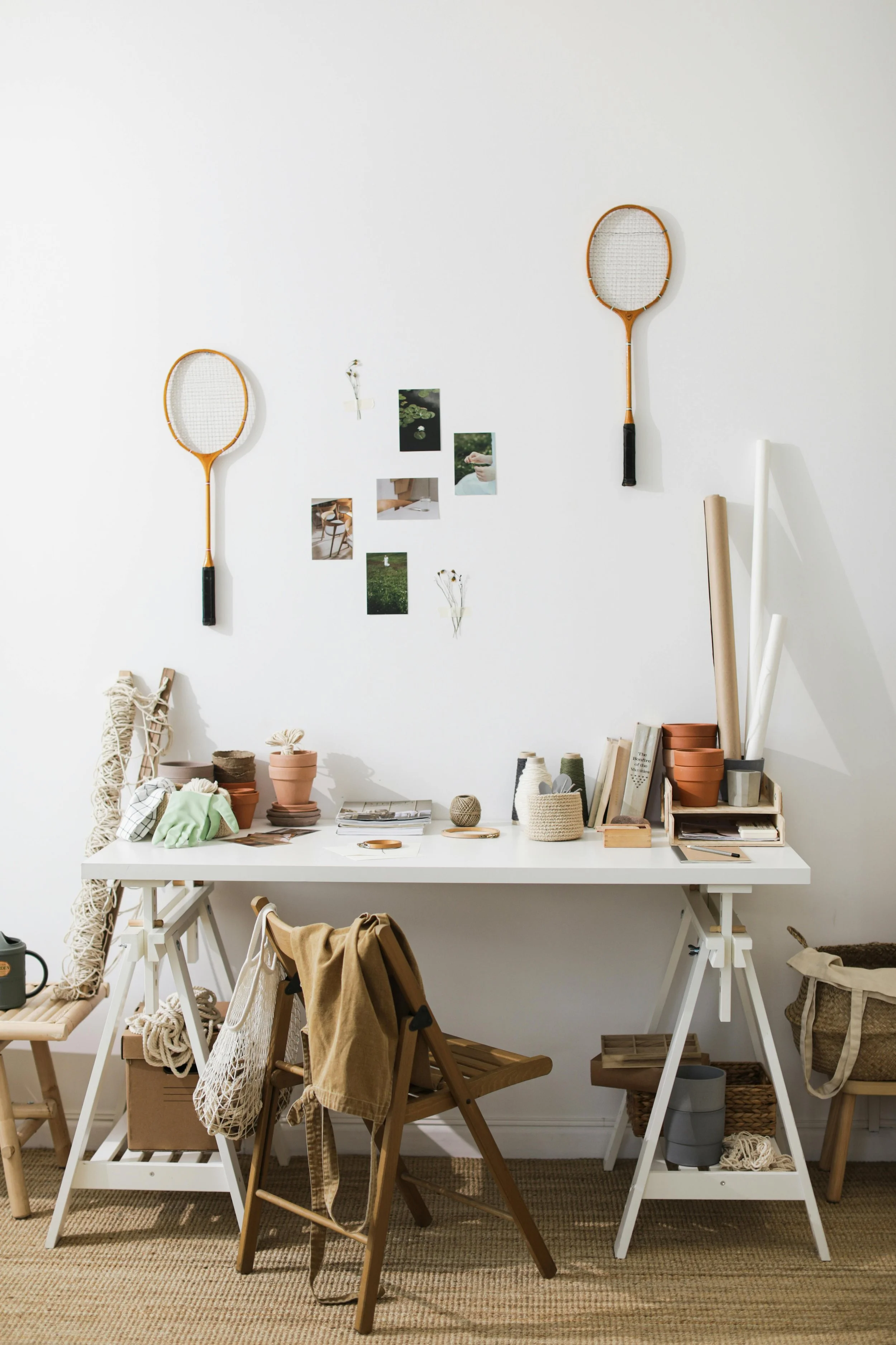 A cozy craft room with a white table, wooden chairs, and various crafting supplies including potted plants, books, and rolls of paper. Two vintage tennis rackets are wall-mounted above, and small photos and dried flowers are decoratively arranged on the wall.