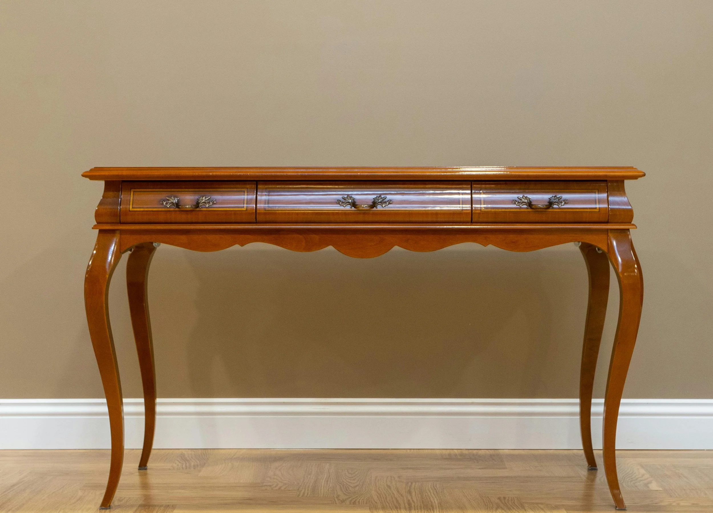 A wooden console table with three drawers, ornate handles, curved legs, and decorative carvings, placed against a beige wall with white baseboards.