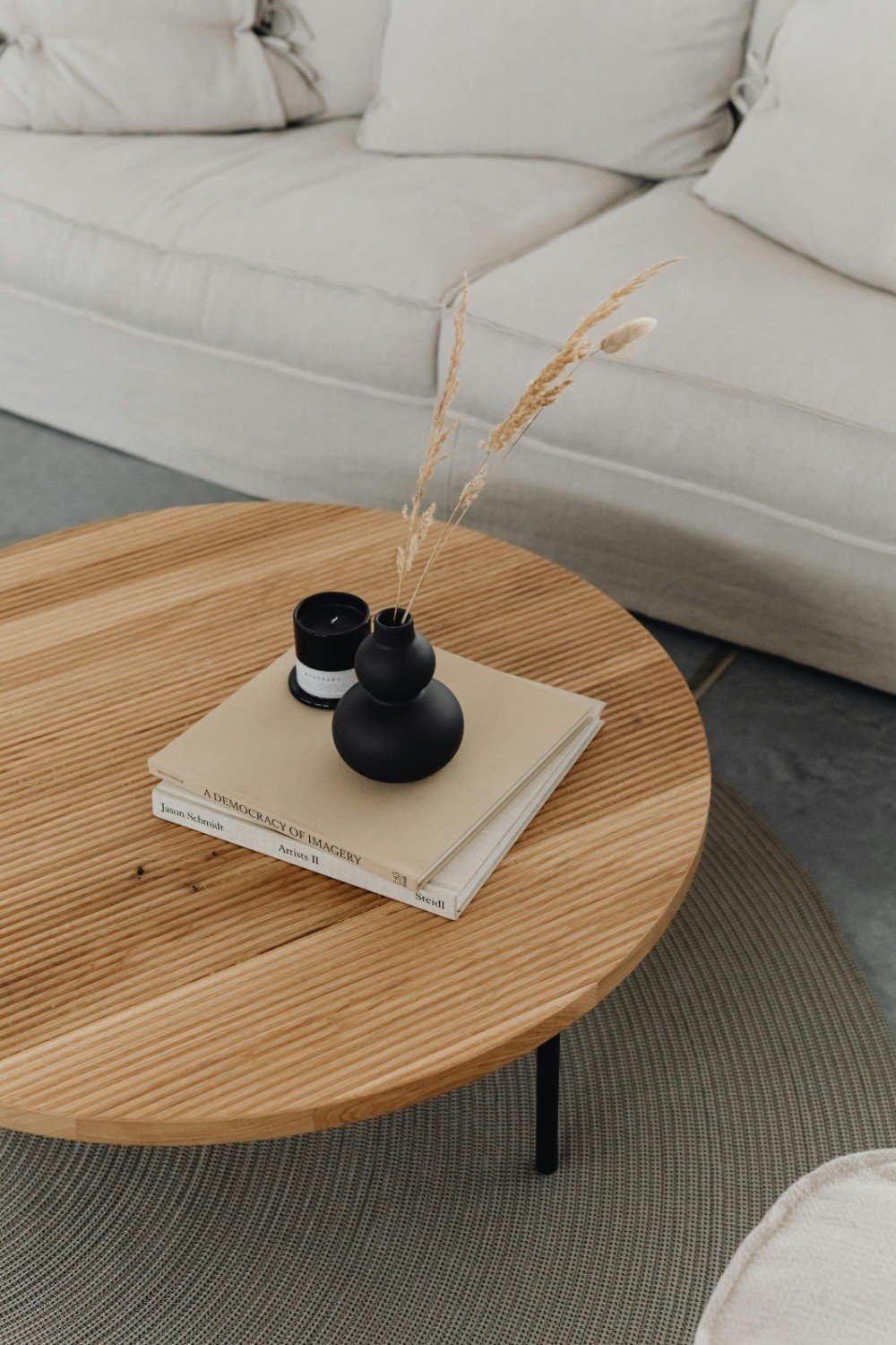 A round wooden coffee table with a black vases with dried grass, a candle, and two books on top, in front of a white sofa.