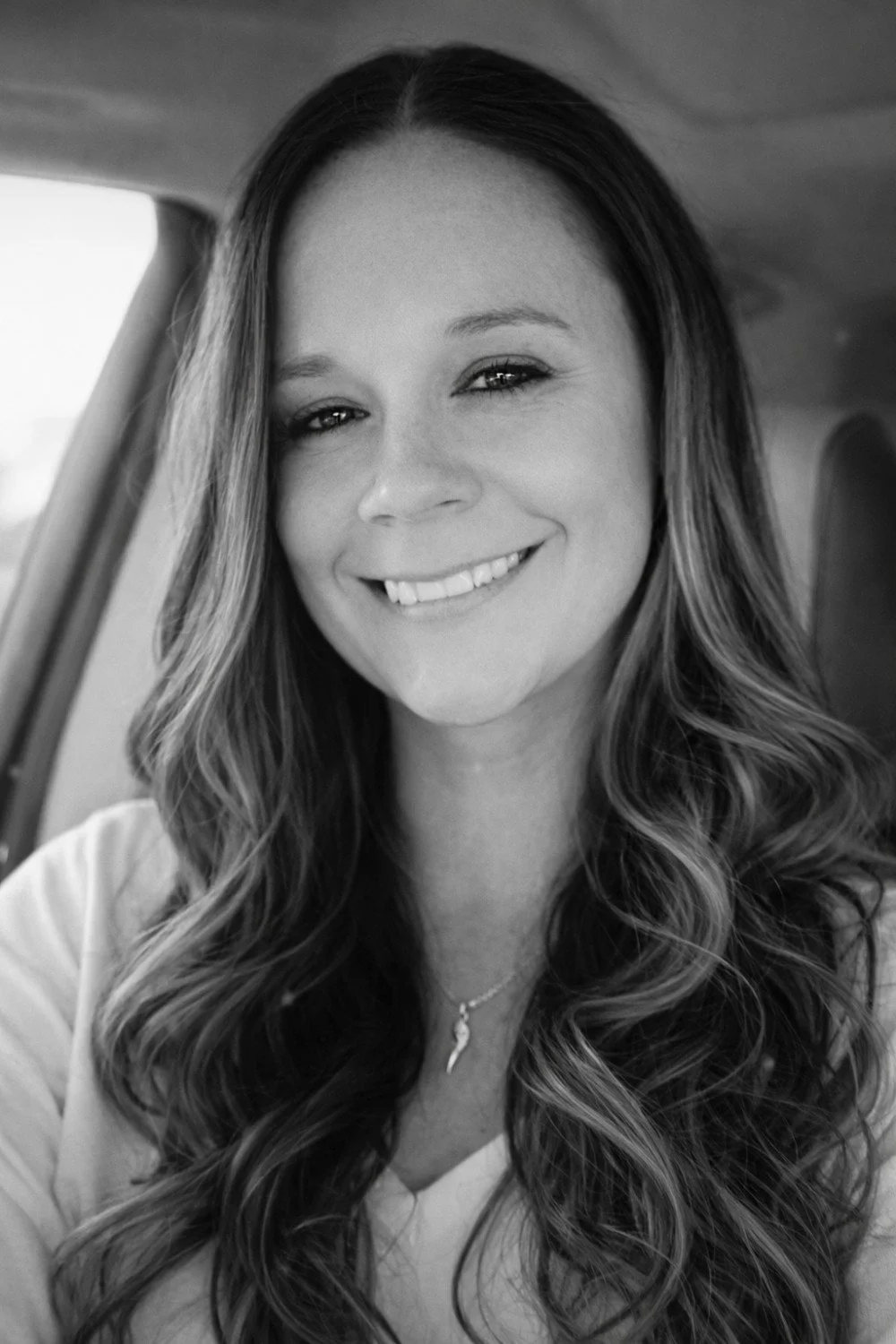 A smiling woman with long wavy hair taking a selfie inside a vehicle.