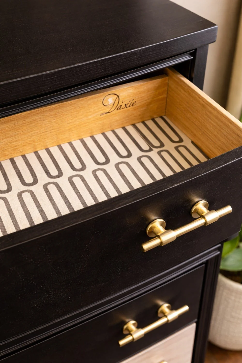 Open black wooden drawer with gold handle, lined with patterned paper and labeled inside with 'Dixie'