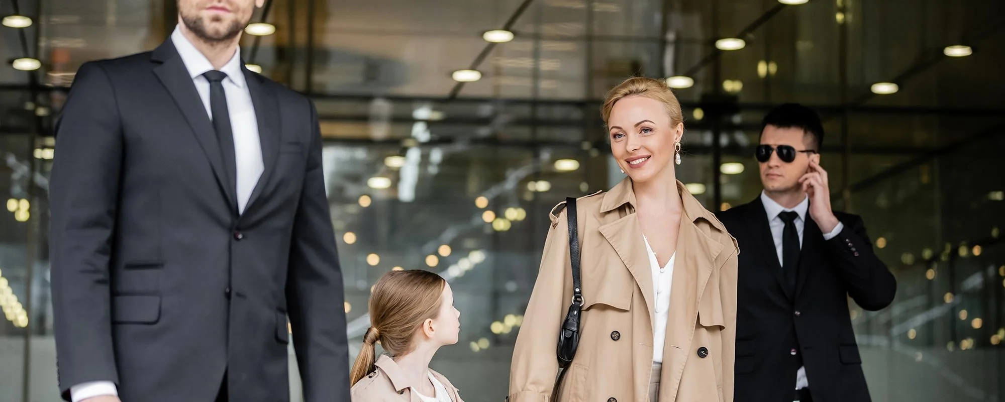 A group of people, including a woman with blonde hair and a young girl, walking through a modern, glass-walled building, with men in suits, one talking on a cell phone, in the background.