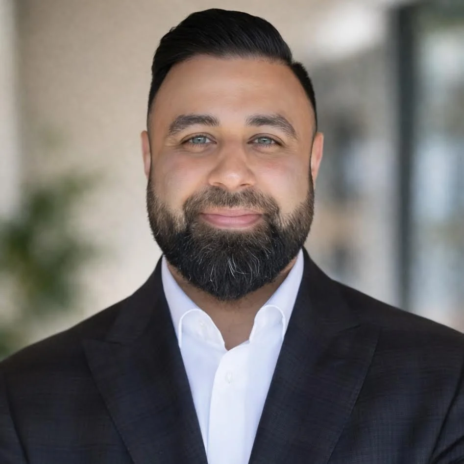Professional man with a beard and styled hair, wearing a black suit and white shirt, smiling confidently.