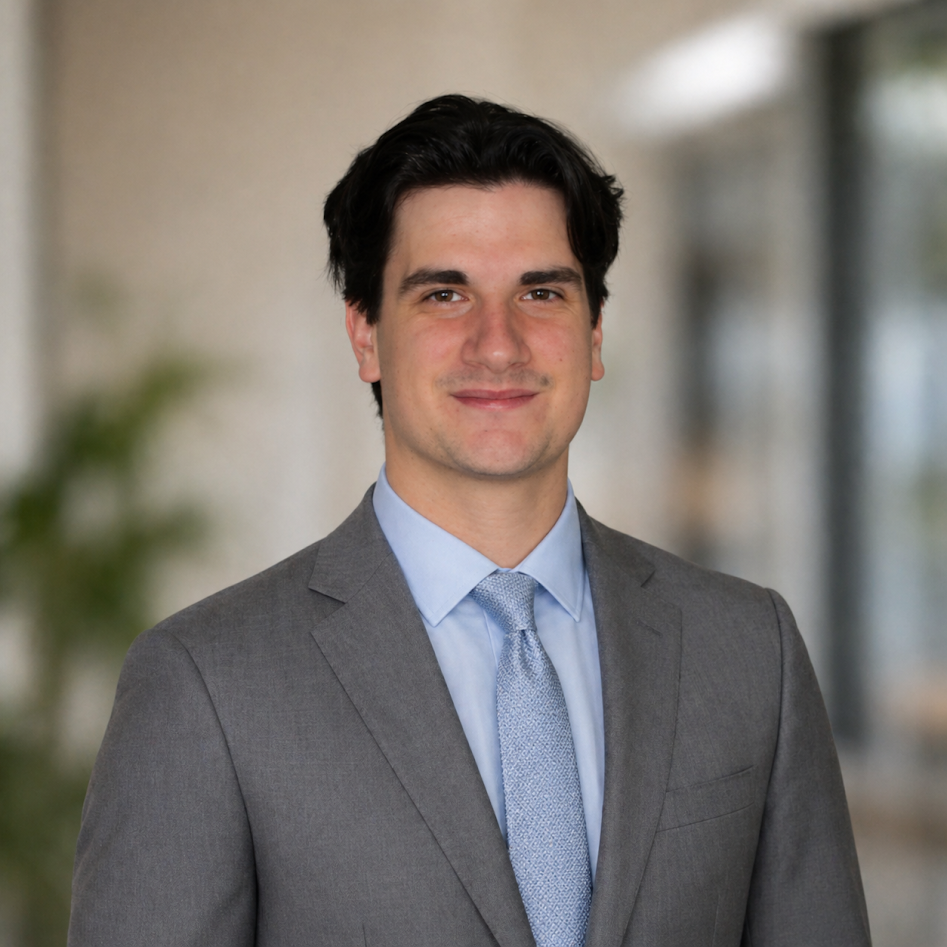 A young man with dark hair wearing a gray suit, light blue shirt, and blue tie, smiling, in an office setting with a blurred background.