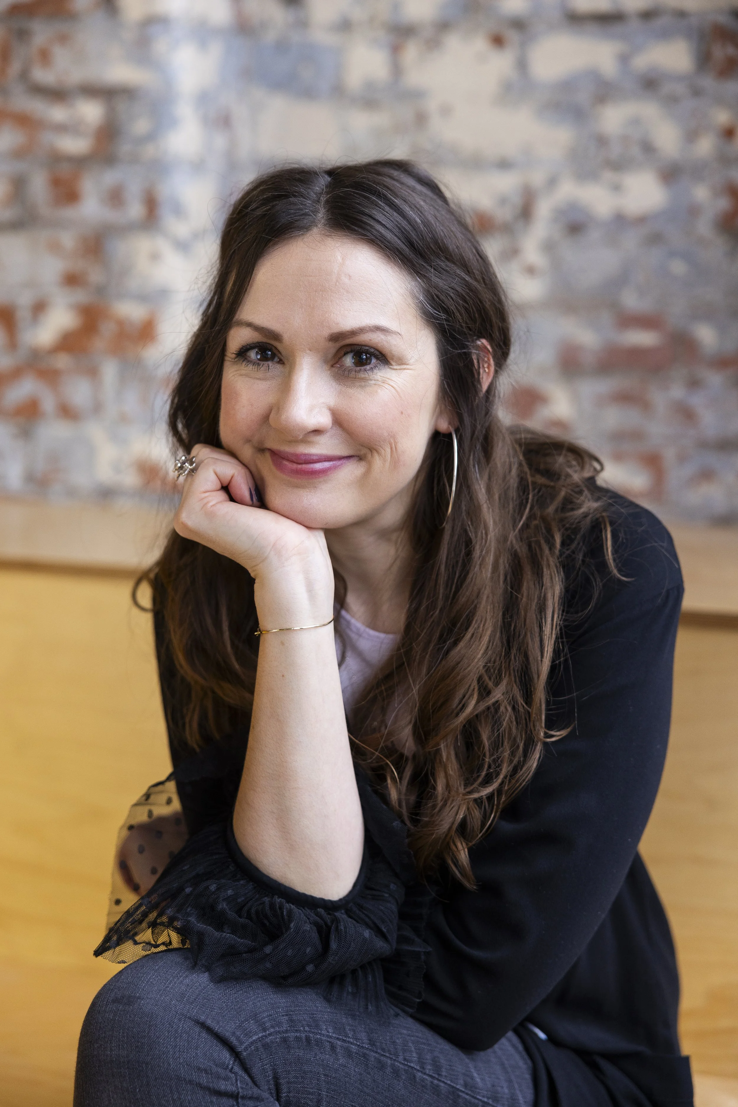 A woman with long brown hair and hoop earrings smiling, resting her chin on her hand, sitting indoors against a brick wall.