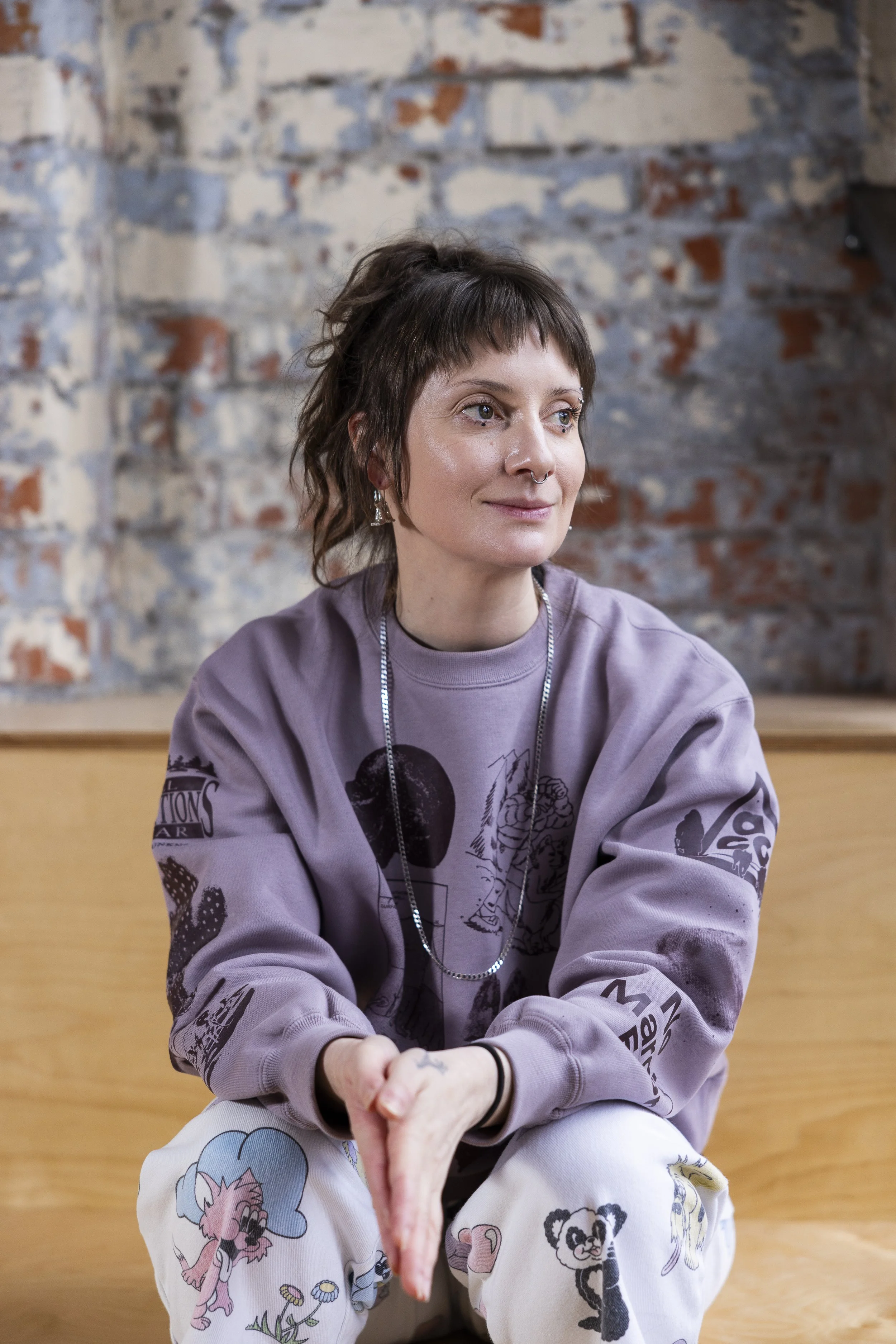 A young woman with short brown hair, wearing a purple sweatshirt and patterned pants, sitting on a wooden surface in front of a brick wall.