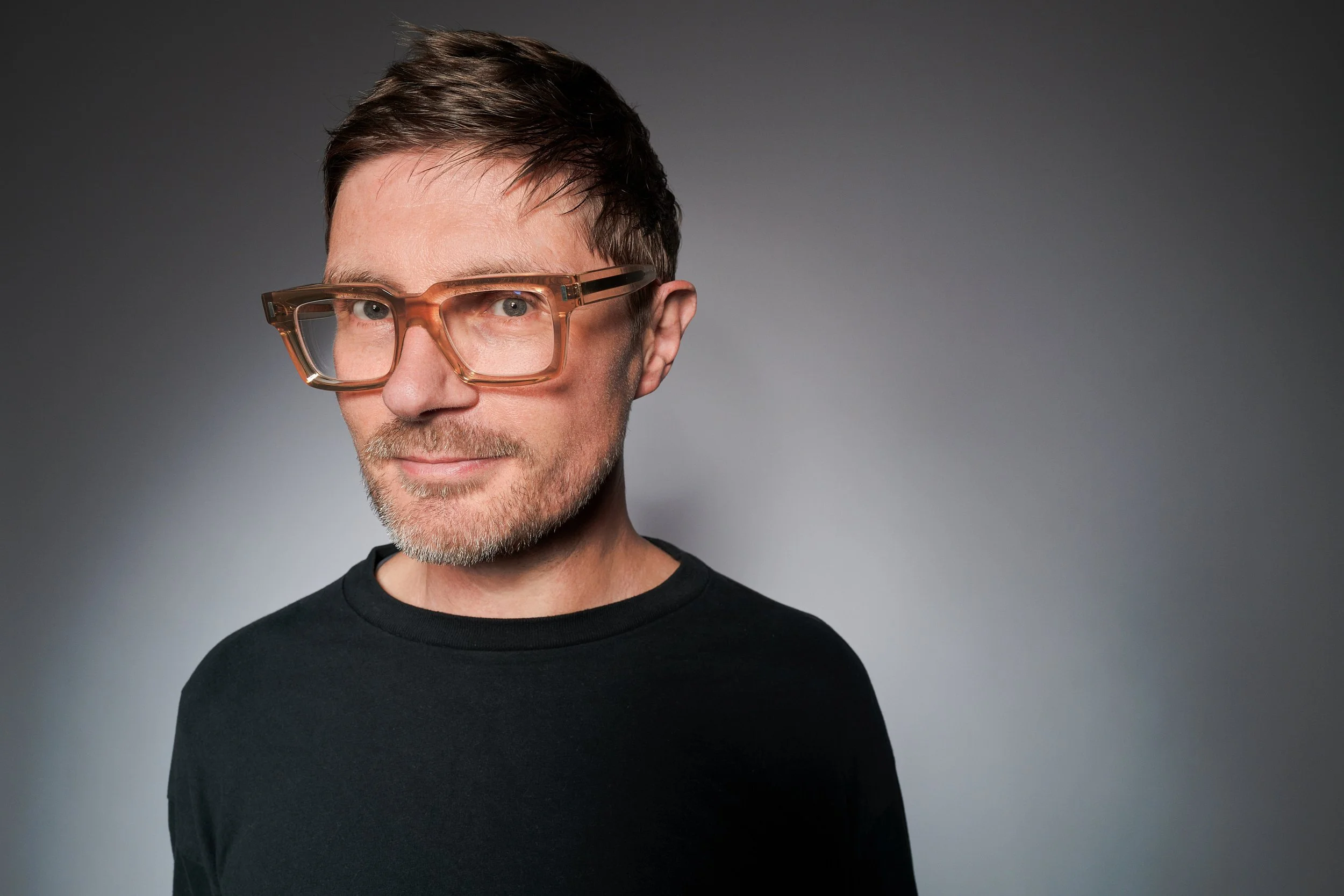 A man with short brown hair, glasses, and a beard wearing a black shirt against a gray background.