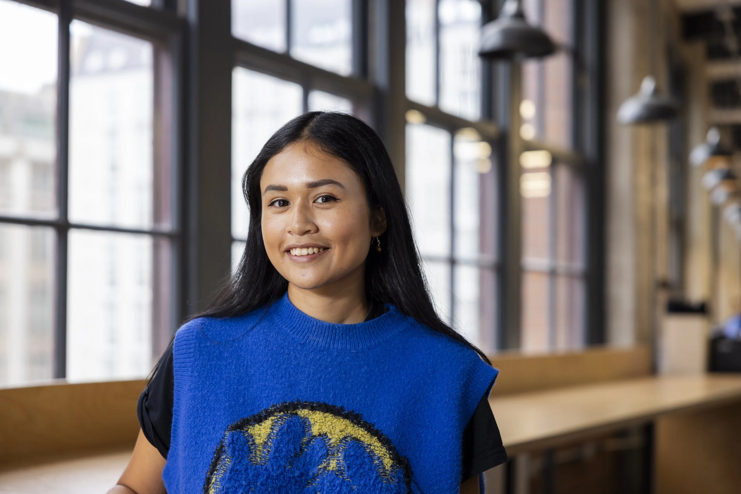 A young woman with long black hair smiling indoors near large windows, wearing a blue sleeveless top with a yellow and black emblem.