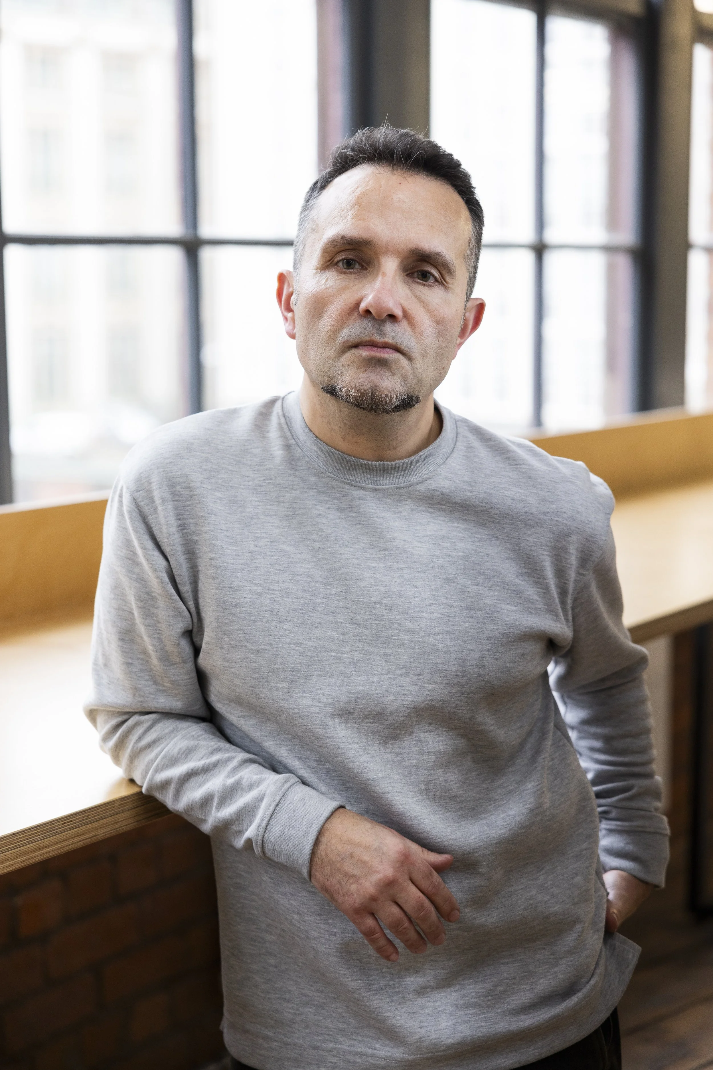 A man with short dark hair, a goatee, wearing a gray sweatshirt, standing indoors near a large window with a city view in the background.